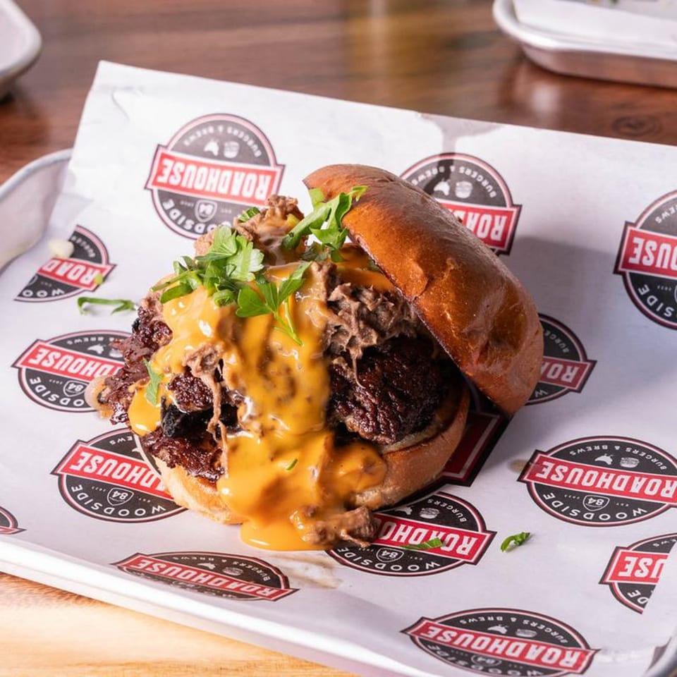 A Woodside Roadhouse pulled pork sandwich with cheese sauce and cilantro on branded paper.